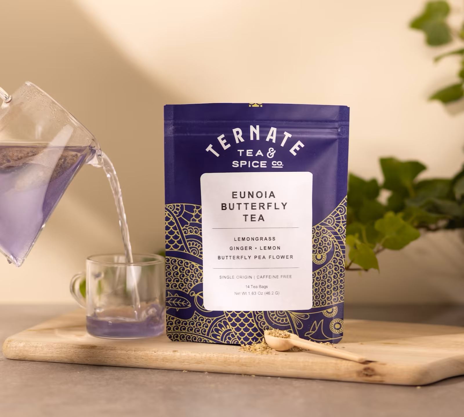 Eunoia Butterfly Tea in navy and gold packaging on a wooden board, with a glass pitcher pouring the brewed violet butterfly pea tea into a clear cup.