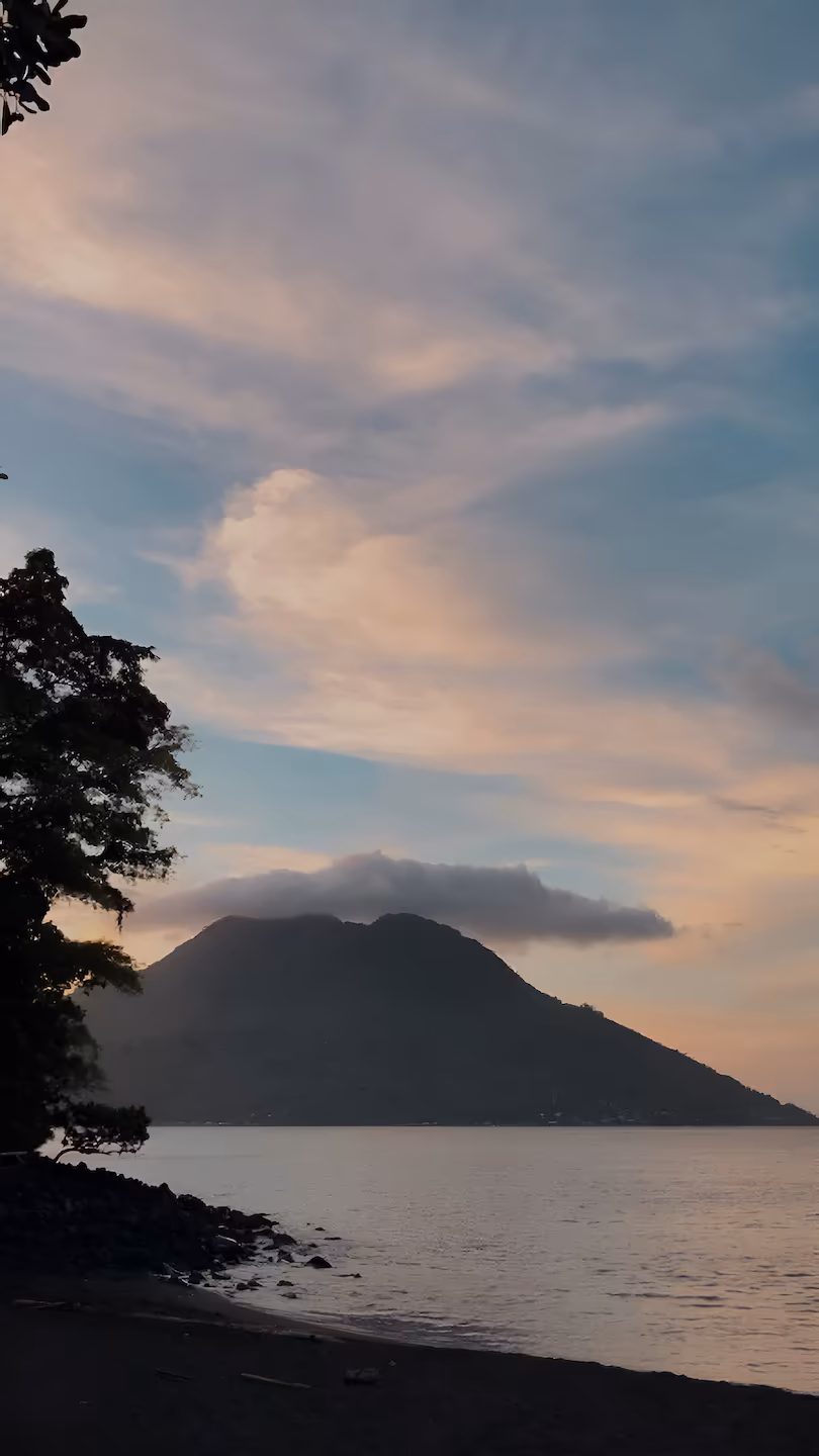 A serene view of Ternate Island’s volcano silhouetted against a soft, pastel-colored sunrise, with calm waters and a tree framing the foreground.