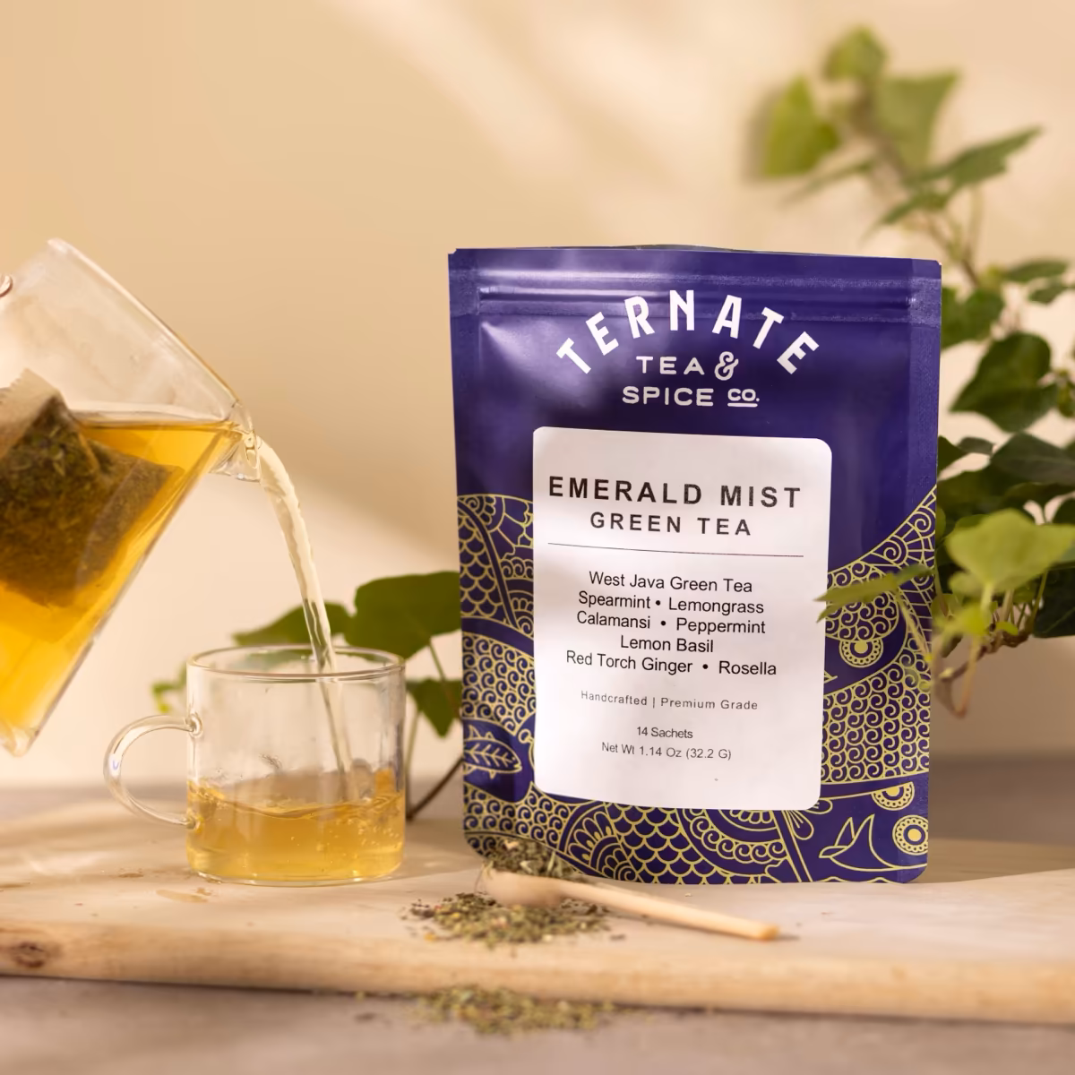 Emerald Mist green tea in navy and gold packaging, with a glass teapot pouring the brewed tea into a clear cup beside loose mint leaves.