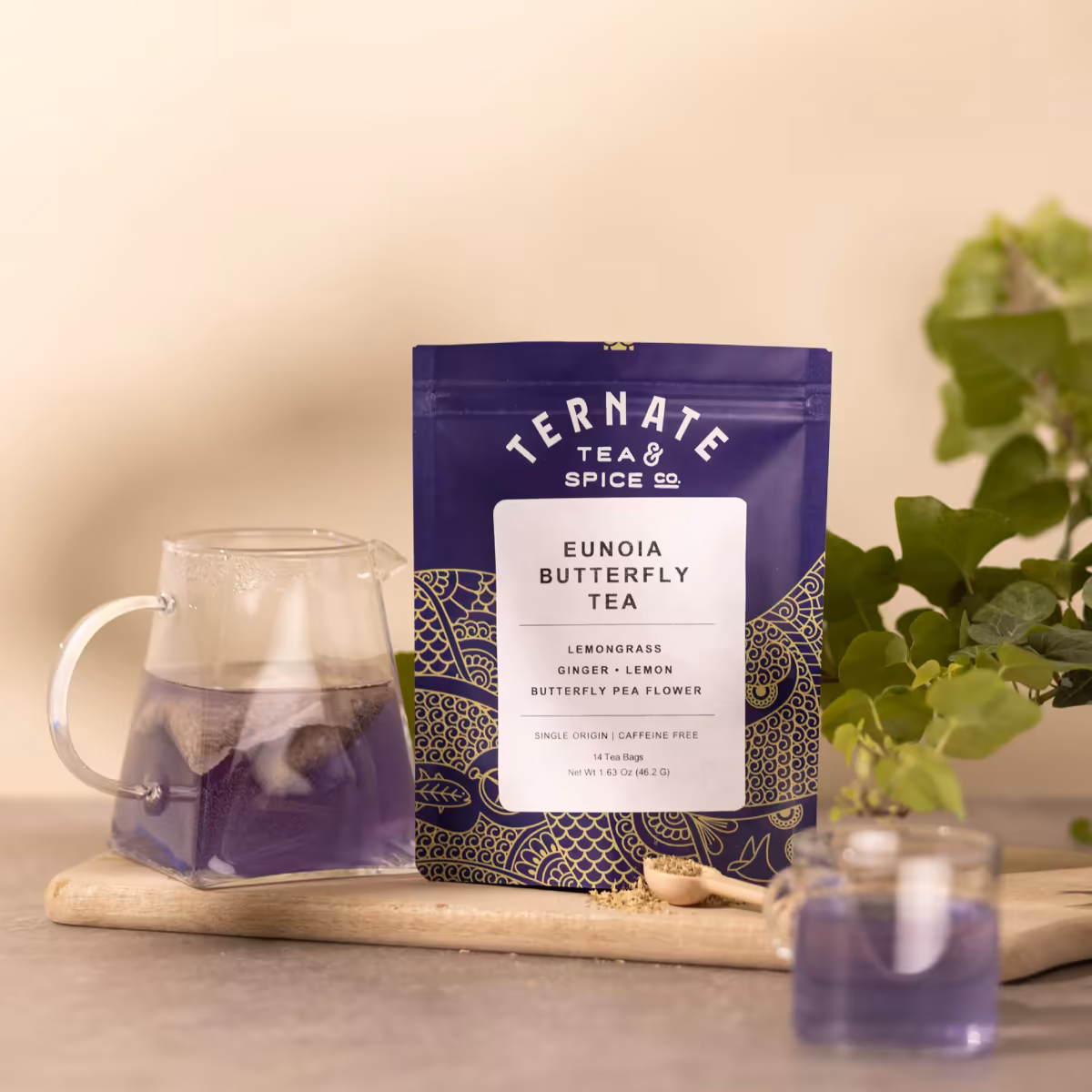 Eunoia Butterfly Tea in navy and gold packaging on a wooden board, beside a glass pitcher and cup of the brewed violet butterfly pea tea.