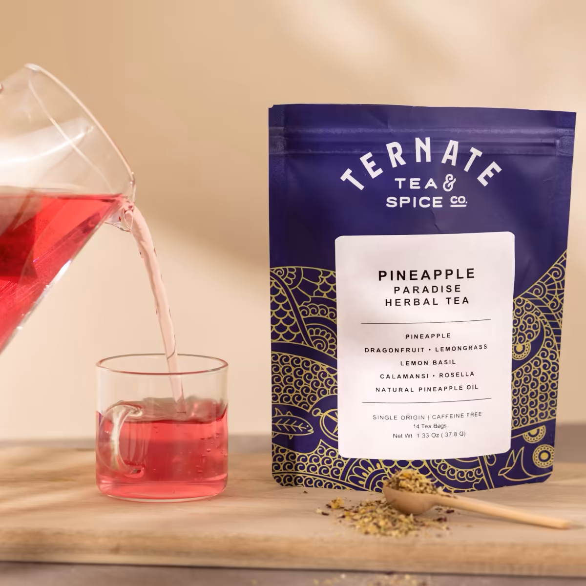 Pineapple Paradise herbal tea in navy and gold packaging, with a glass pitcher pouring the brewed rose-pink tea into a clear cup.